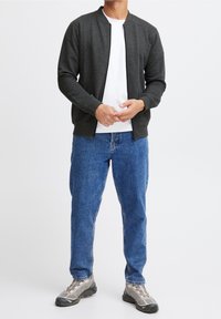 Ciemnoszara kurtka bomber z zamkiem błyskawicznym, z prążkowanymi mankietami i dolnym wykończeniem, noszona na białym t-shircie, w połączeniu z niebieskimi dżinsami i szarymi trampkami.