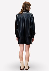 Zwarte nepleren oversized blouse met een samengevoegde achternaad, gecombineerd met bijpassende shorts. Model draagt lichtcrème sokken en donkere sandalen.