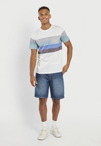 Kortærmet hvid t-shirt med horisontale striber i grå, blå og teal, parret med blå denim shorts og hvide sneakers.