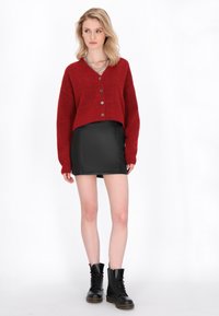 Rote kurz geschnittene Strickjacke mit fünf Knöpfen, kombiniert mit einem schwarzen Minirock aus Kunstleder und schwarzen Schnürkampfstiefeln. Glatte Textur überall.