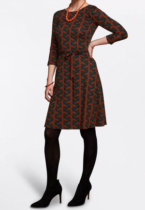 Kobieta ubrana w zielono-pomarańczową, wzorzystą sukienkę długości do kolan, czarne rajstopy, czarne szpilki i pomarańczowy naszyjnik z koralików, stojąca z jedną ręką na biodrze.