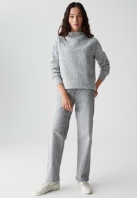 Helles graues Fleece-Pullover mit hohem Kragen, kombiniert mit hellgrauen geraden Jeans und weißen Sneakern. Das Model steht vor einem schlichten Hintergrund.