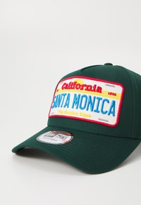 Căciulă de baseball verde, decorată cu un patch brodat pe care se află textele "California", "SANTA MONICA" și "The Golden State" în nuanțe de albastru, roșu și galben.