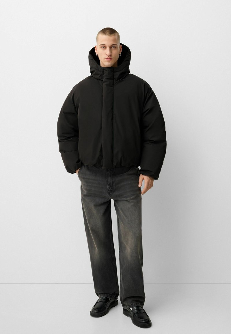 Parka nera con collo alto e cappuccio, caratterizzata da una texture liscia e lucida. Indossata con jeans ampi grigio scuro sbiaditi e scarpe nere.