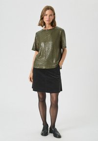 Top scurt cu mânecă, de culoare verde măsliniu, cu paiete, asortat cu o fustă neagră; modelul poartă pantofi negri și ciorapi negri subțiri, stând în fața unui fundal simplu.