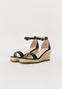 Sandalias de cuña de cuero negro con suela de yute estilo espadrille, punta abierta y tira en el tobillo con detalle de hebilla. Textura suave, diseño casual.