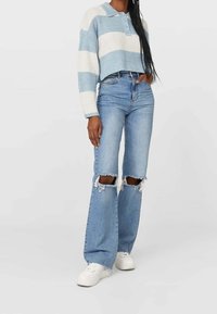 Donna che indossa un maglione a righe blu chiaro e bianche, jeans blu strappati a vita alta e sneaker bianche, in piedi davanti a uno sfondo uniforme.