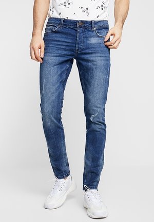 Homme portant un jean slim bleu avec légère usure et des baskets blanches, debout devant un fond uni.