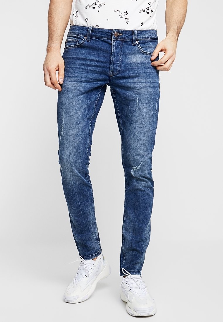 Homme portant un jean slim bleu avec légère usure et des baskets blanches, debout devant un fond uni.