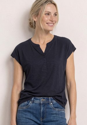 Kvinde iført en mørkeblå henley med korte ærmer, stukket ned i blå jeans, stående og smilende mens hun kigger til siden.