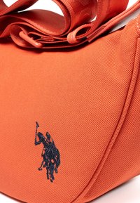 Orangene, strukturierte Stofftasche mit einem dunkel gestickten Logo, das einen Reiter zeigt. Verfügt über einen breiten Träger und eine runde Form.