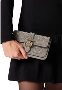 Clutch purse metallica grigia con una superficie texturizzata, hardware dorato e una chiusura decorativa a anello. Indossata a mano, abbinata a un vestito nero.