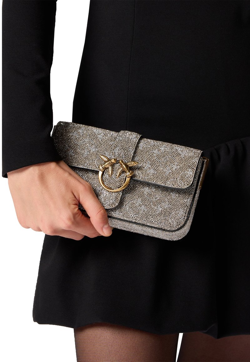 Clutch purse metallica grigia con una superficie texturizzata, hardware dorato e una chiusura decorativa a anello. Indossata a mano, abbinata a un vestito nero.