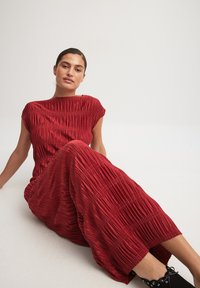Red pleated dress with a boat neckline and short sleeves, featuring a soft texture and a full skirt. Black shoes with laces complete the look.