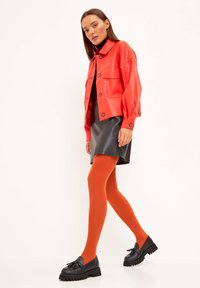 Giacca cropped rossa in pelle con bottoni, abbinata a una gonna nera in ecopelle, collant arancioni e scarpe platform nere con fiocchi.