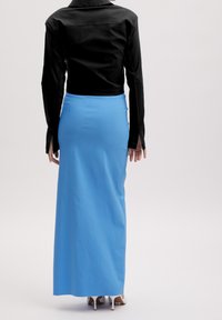 Person, der mit dem Rücken zur Kamera steht und ein schwarzes Langarmshirt, einen blauen Maxikleid und weiße High Heels auf einem einfarbigen Hintergrund trägt.