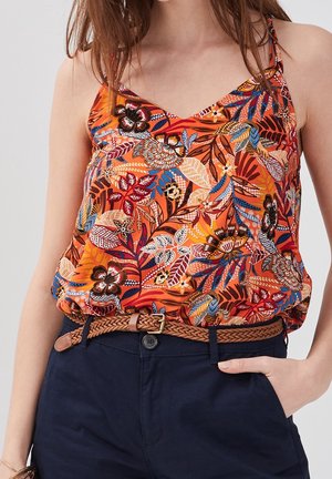 Femme portant un haut sans manches orange à motifs floraux, rentré dans un pantalon bleu marine avec une ceinture tressée marron, main dans la poche, cheveux lâchés.