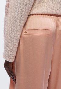 Pantalon en satin rose pâle à la texture lisse, avec une poche arrière et un petit logo doré. Stylisé avec un pull crémeux duveteux.