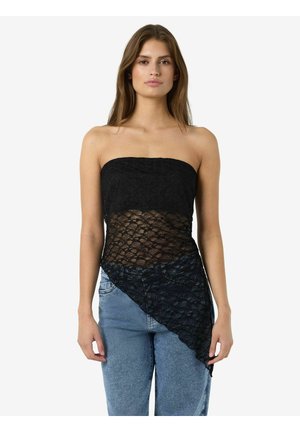 Femme portant un top bustier en dentelle noire avec un ourlet asymétrique sur un jean bleu, debout devant un fond blanc uni.