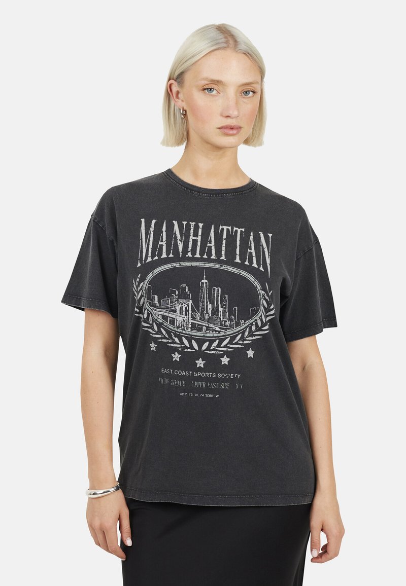 Femme aux cheveux blonds courts portant un t-shirt noir surdimensionné avec le texte blanc "Manhattan" et un graphique de la silhouette de la ville.