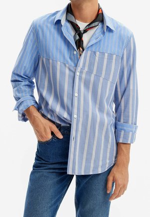 Mand iført en blå og hvid, vertikalt striped skjorte med knapper, med ærmerne rullet op, mørkeblå jeans og et sort og orange tørklæde.
