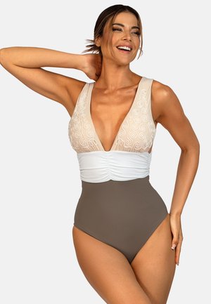 Uśmiechnięta kobieta w bezrękawnym stroju kąpielowym z głębokim dekoltem w kształcie litery V, z beżowym haftowanym topem, białym środkiem i brązowymi majtkami z wysokim stanem, stoi z ręką założoną za głowę.