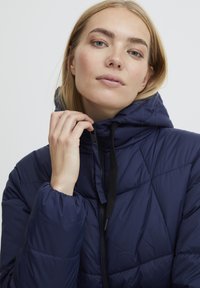 Marineblaue Daunenjacke mit Kapuze, ausgestattet mit einem rautenförmigen Steppdesign, Reißverschluss vorne und elastischen Bündchen. Glatte, glänzende Textur.