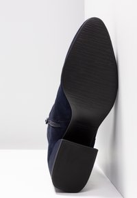 Bottine en daim bleu foncé avec semelle noire texturée, fermeture éclair sur le côté et talon carré épais vue du côté de la semelle.