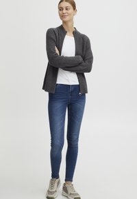Mørkegrå ribbestrikket cardigan med høy krage, hvit t-skjorte under, blå smale jeans og beige joggesko med grå detaljer.
