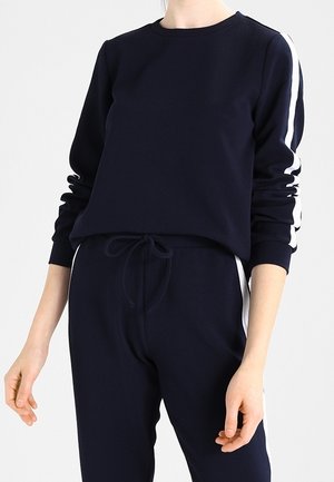 Felpa navy con maniche lunghe, collo tondo e strisce bianche sui lati. Il tessuto è liscio con una vestibilità comoda, abbinata a pantaloni jogger coordinati.
