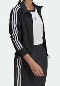 Svart sportjacka med vita ränder på ärmarna, tillsammans med en vit t-shirt med en liten logotyp. Slät tyg, frontdragkedja.