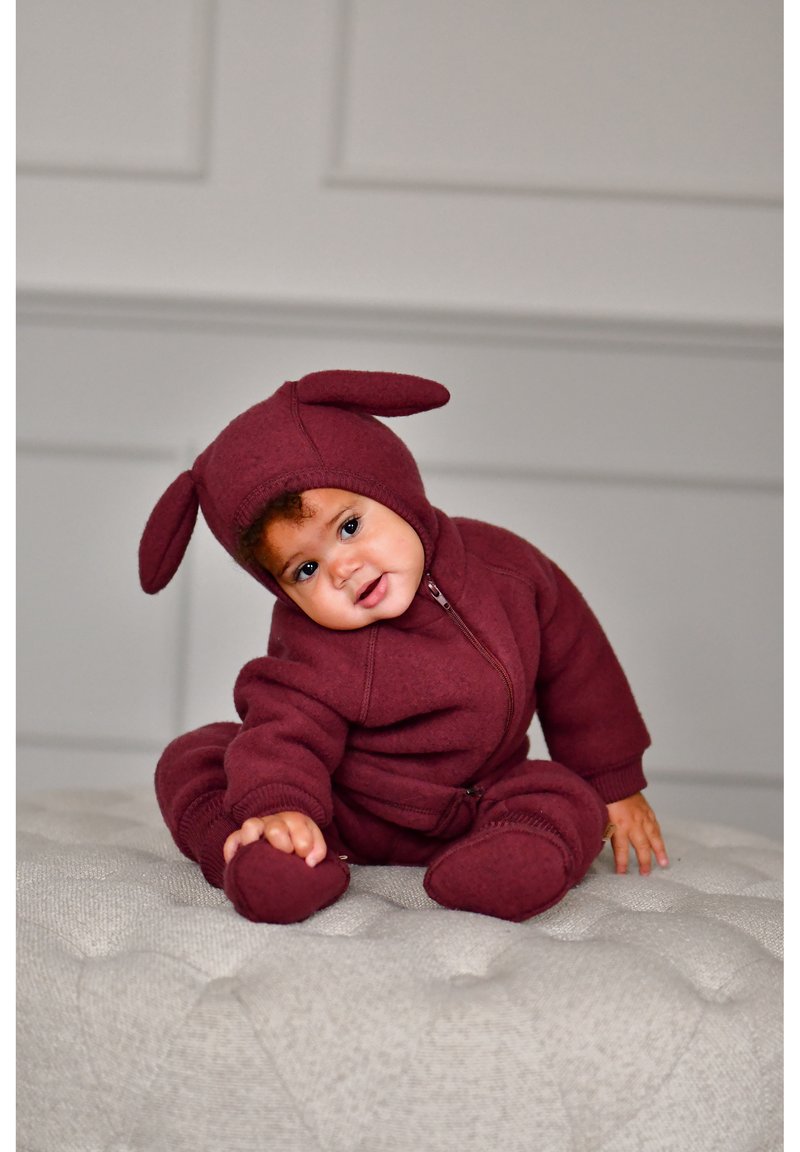 Bordový overal z fleecu na zips s králičími uškami, príjemnou textúrou, rebrovanými detailmi a elastickými manžetami, navrhnutý pre dojčatá, sediaci na svetlom povrchu.