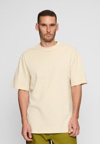 Beige oversized póló rövid ujjakkal és magas nyakkal, sima pamutból készült, laza szabású és minimális varrásokkal.