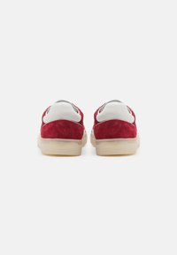 Paire de baskets rouges en suède avec des languettes de talon en cuir blanc et des semelles en caoutchouc beige translucide, vues de dos sur fond blanc.