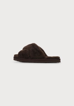 Zapatilla marrón oscuro y peluda con diseño cruzado y suela plana sobre un fondo claro y liso.