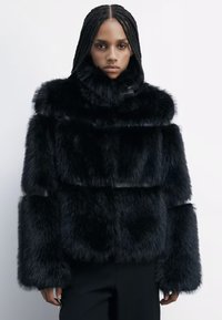 Veste en fausse fourrure noire avec un col haut, un design volumineux et des manches larges. Présente des panneaux texturés et une apparence chunky et douce.