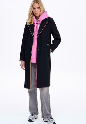 Blondynka w czarnym długim płaszczu z kolorowymi wykończeniami, nosząca różowy sweatshirt, szare dżinsy i białe sneakersy, stoi z jedną ręką w kieszeni.