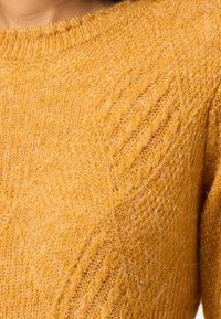 Musztardowożółty dziergany sweter z fakturowanym wzorem i prążkowanym dekoltem. Miękki materiał wełniany o luźnym kroju i długich rękawach.