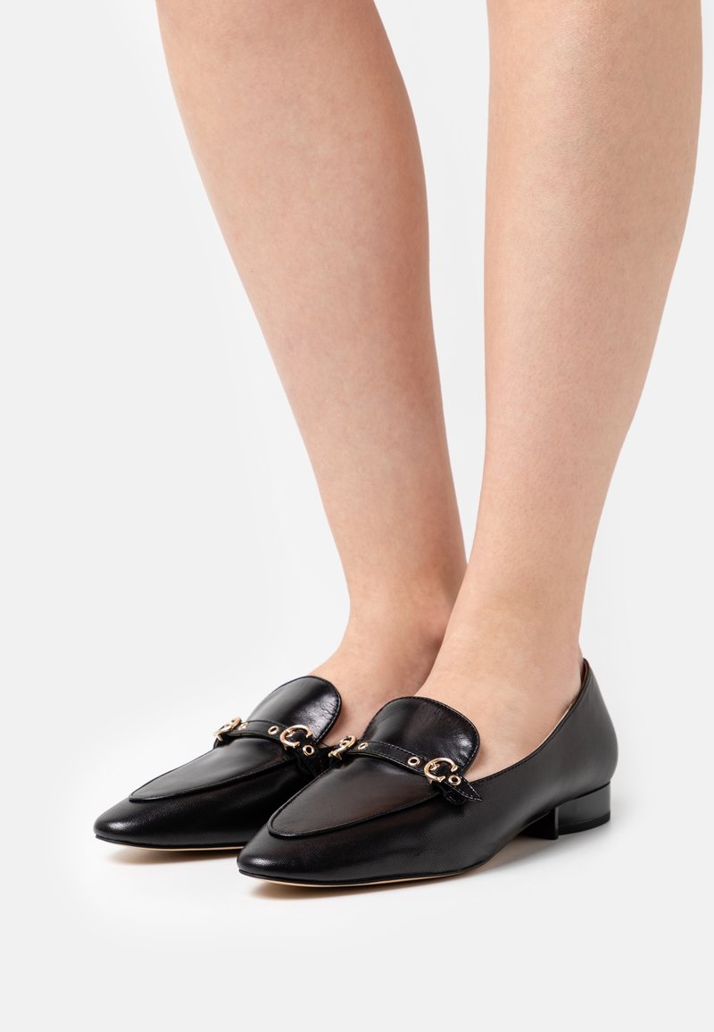 Mocassins en cuir noir avec des détails de boucle dorée portés pieds nus sur fond blanc.