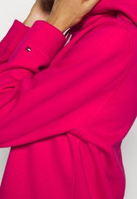 Bluza z kapturem w kolorze fuksji z regulowanym sznurkiem i ściągaczami na rękawach. Posiada małe logo na rękawie oraz miękką, teksturowaną tkaninę.