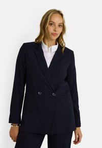 Blazer de corte cruzado azul marino con rayas, con solapas de muesca, dos bolsillos frontales y detalles de botones. Llevado sobre una camisa blanca con cuello.
