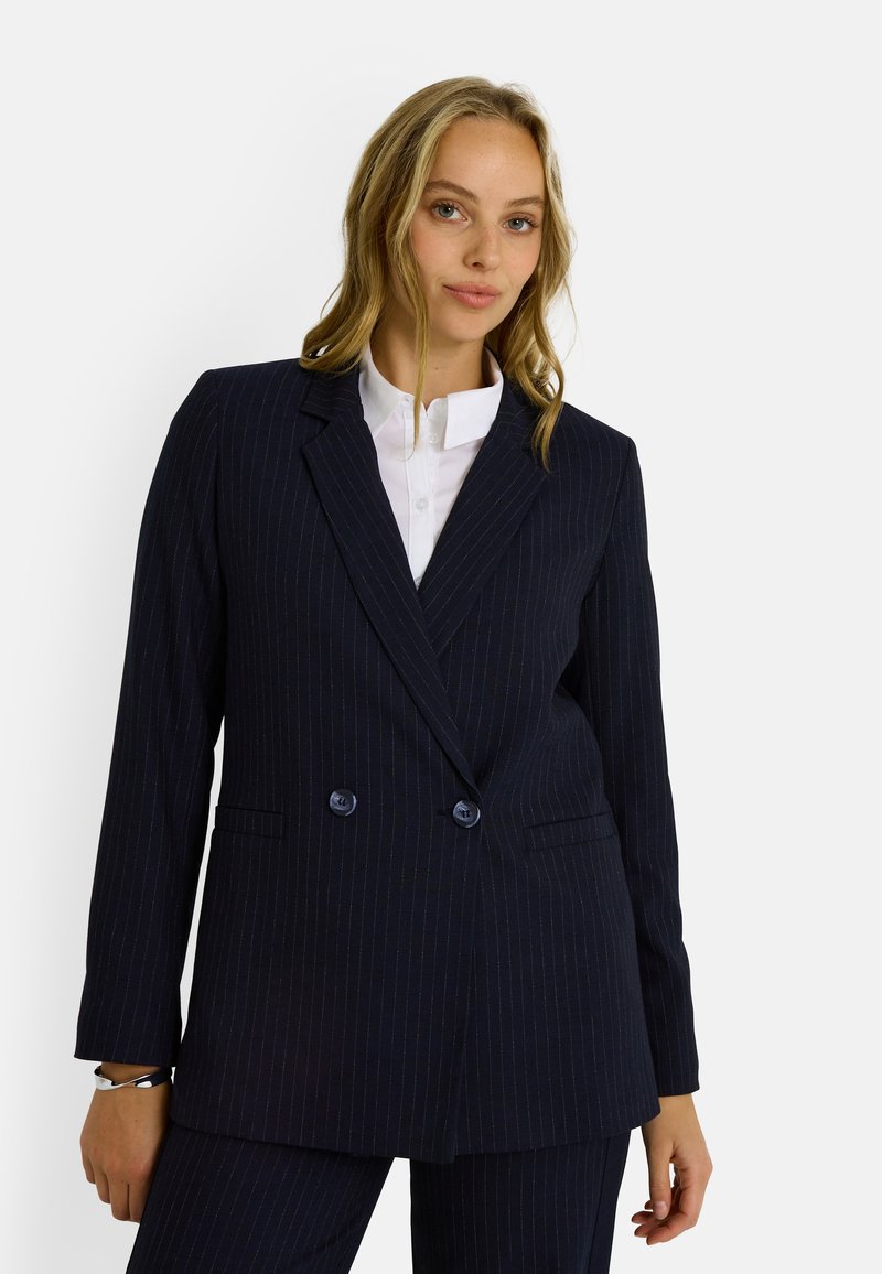 Blazer de corte cruzado azul marino con rayas, con solapas de muesca, dos bolsillos frontales y detalles de botones. Llevado sobre una camisa blanca con cuello.