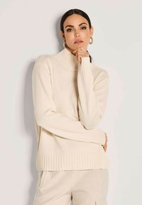 Frau mit dunklem Haar, die einen cremefarbenen Rollkragenpullover und passende Hose trägt, posiert mit der Hand nahe am Kinn vor einem hellen Hintergrund.