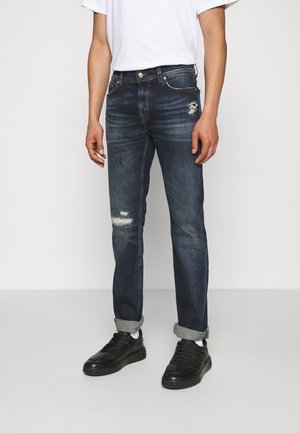 Mand iført mørkeblå slim-fit jeans med slidmærker på låret og knæet, hvide strømper, sorte sneakers og en hvid t-shirt.