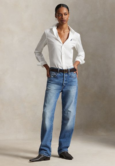 Mujer de pie con las manos en los bolsillos, vistiendo una camisa blanca abotonada, jeans azules, cinturón negro y botas negras puntiagudas, contra un fondo neutro.