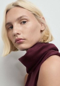Mujer rubia con cabello corto que lleva un suéter sin mangas de color marrón, mirando ligeramente por encima de su hombro contra un fondo liso.