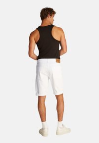 Pantalones cortos de mezclilla blancos con dobladillos enrollados, combinados con una camiseta sin mangas negra. Los pantalones cortos tienen un parche de logo marrón en la cinturilla trasera. Se llevan zapatillas blancas.