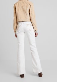 Veste en velours côtelé beige associée à un jean évasé blanc avec des poches brodées. La veste a un design cintré et des détails de boutons.