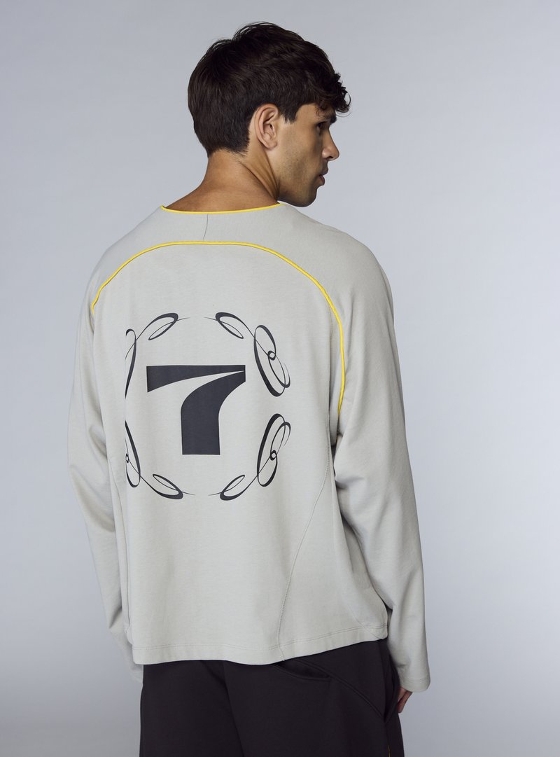 Felpa grigia chiaro con maniche lunghe, presenta un grande numero 7 nero sul retro, evidenziato da cuciture gialle lungo le cuciture.