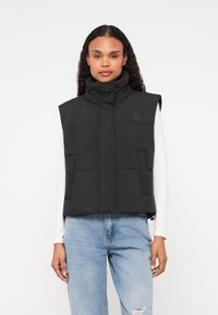 Μαύρο επενδυμένο γιλέκο με ψηλό γιακά, διαθέτει λογότυπο, δύο μπροστινές τσέπες και συντομευμένο σχέδιο. Φοριέται πάνω από ένα λευκό μπλουζάκι και τζιν παντελόνι.